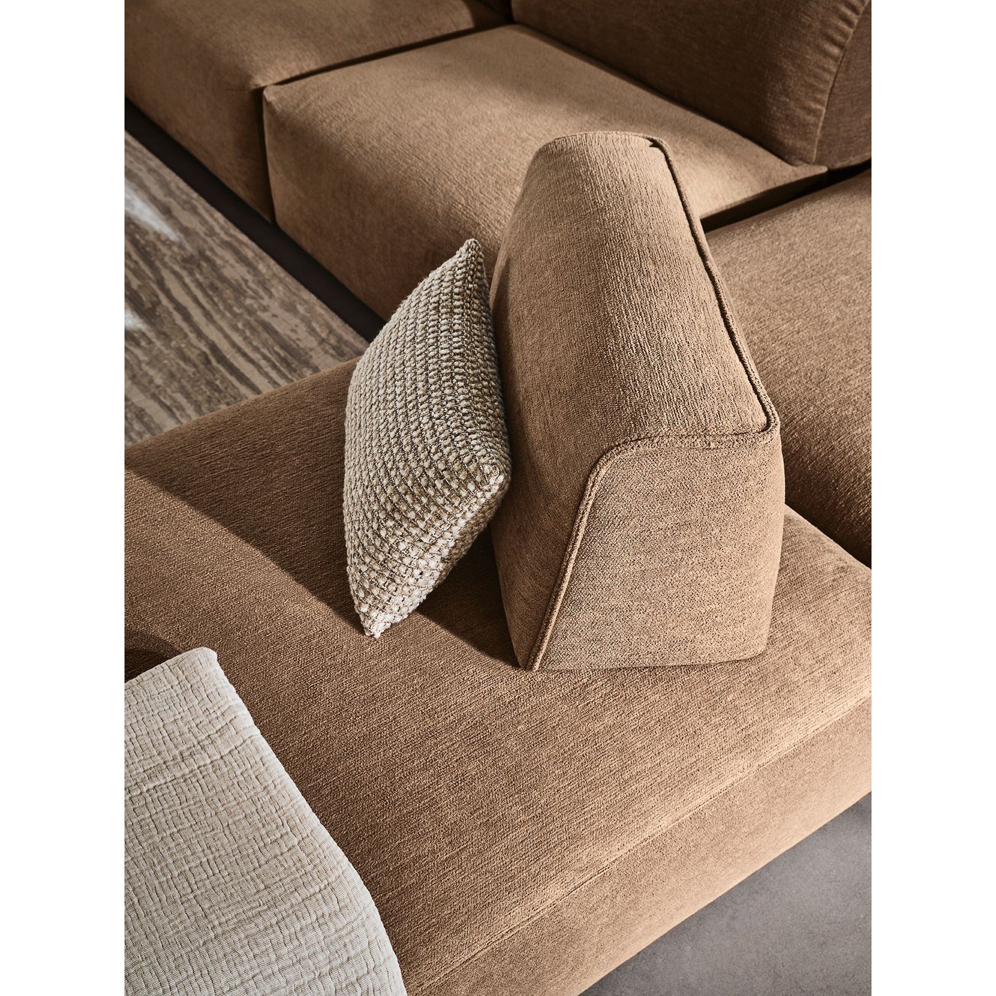 A close-up of a modern sectional sofa in a warm taupe-brown textured fabric, styled with two accent pillows including one chunky knit throw pillow.