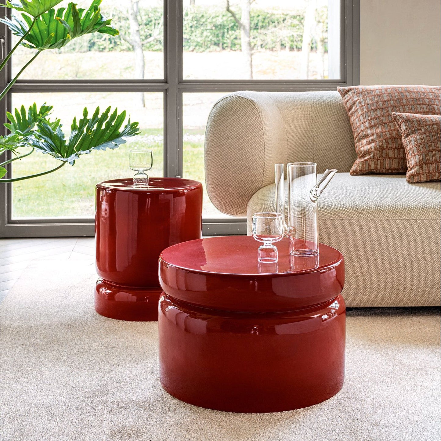 Two glossy deep red round nesting side tables in a modern living room setting with a beige sofa, window view, and green plant.