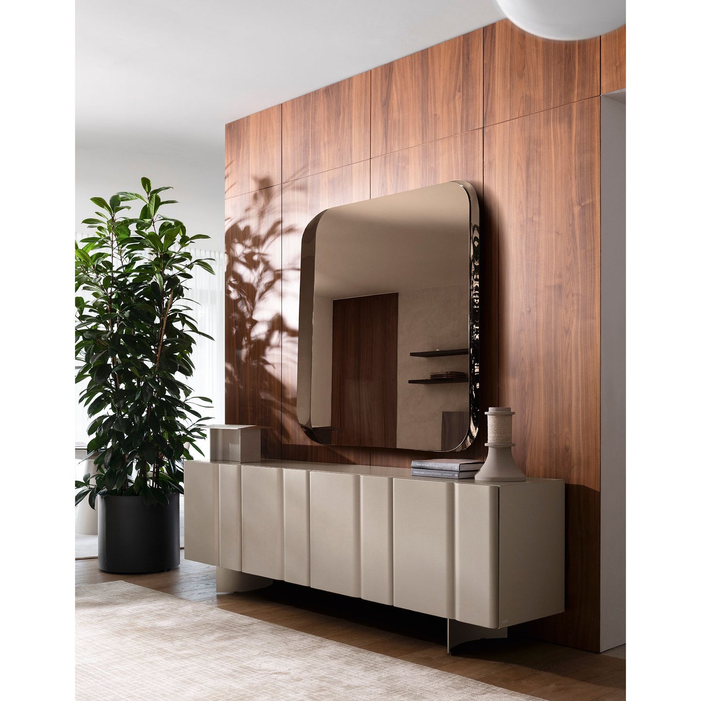A modern sideboard in a warm beige tone with a large arched mirror mounted above it, set against a wood-paneled wall in a styled interior. A potted plant and decorative objects are visible on and around the cabinet.