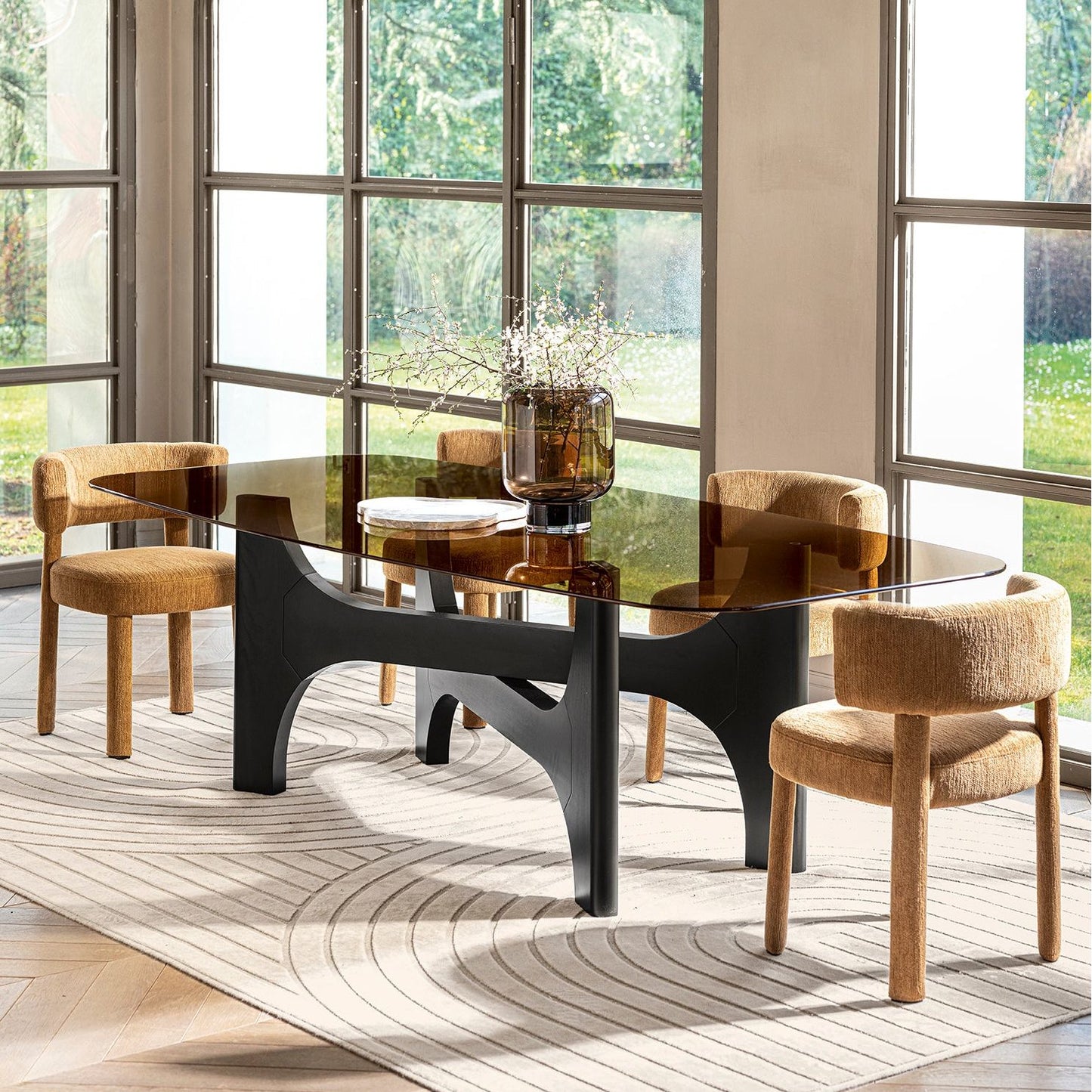 A modern dining set featuring a rectangular table with a dark smoked glass top and a sculptural black metal base, surrounded by four upholstered dining chairs in a warm beige fabric. The set is styled on a patterned cream rug in a bright room with large arched windows looking out onto greenery.