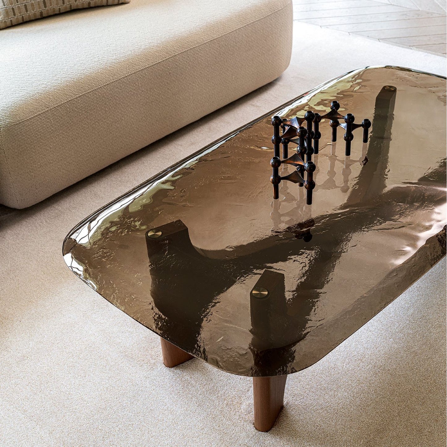 A modern rectangular coffee table with a glossy, hammered metallic bronze-brown top and wooden legs, styled in a minimalist living room next to a beige sofa.