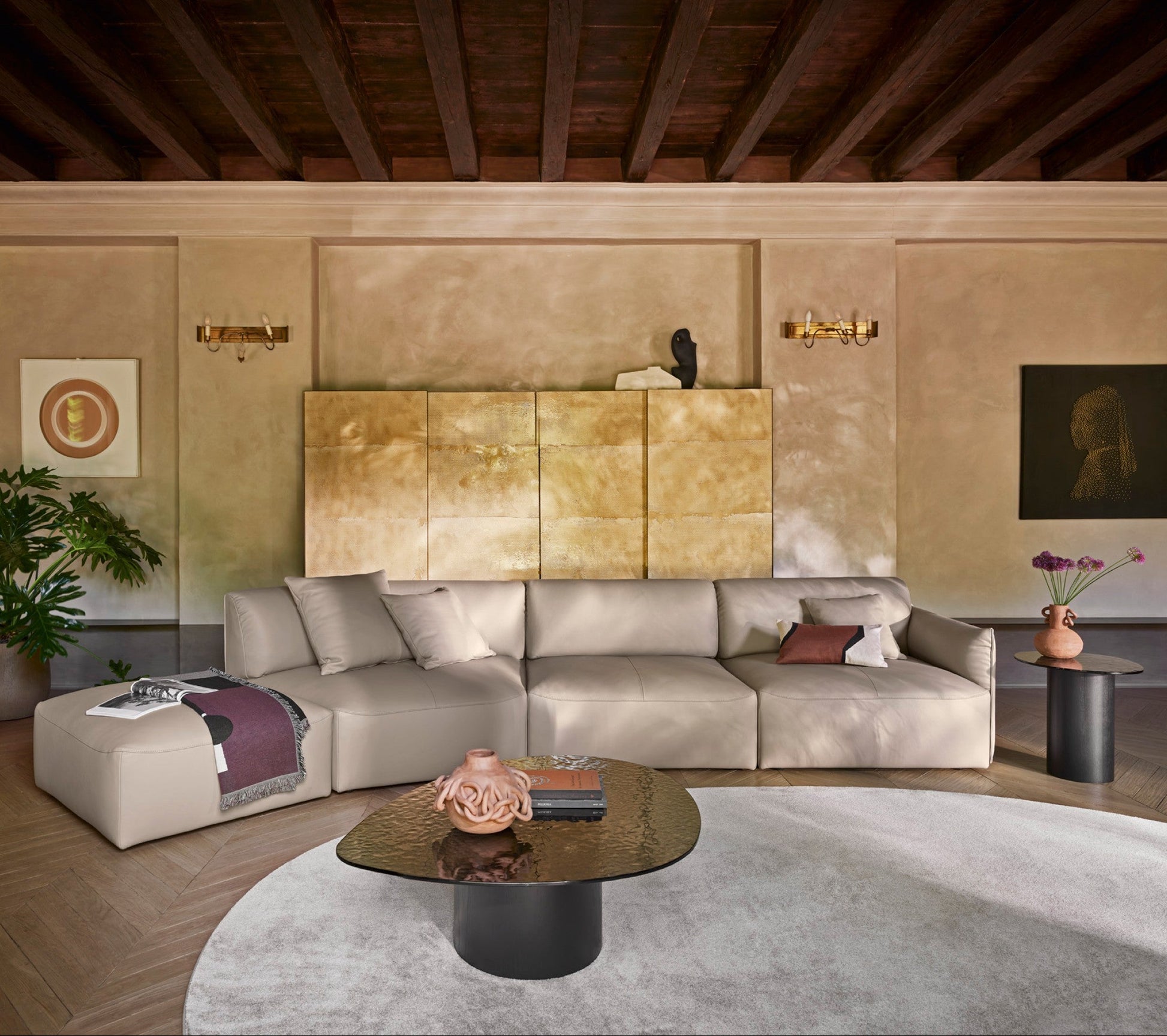 A modern beige sectional sofa with matching ottoman in a styled living room, featuring a round glass-top coffee table, textured beige walls, a large gold-toned panel, and dark wood ceiling beams.