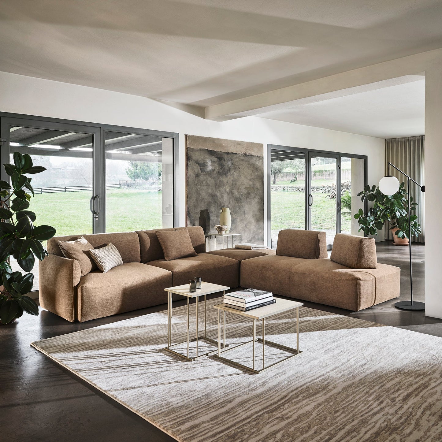 A modern sectional sofa in a warm taupe-brown color arranged on a large textured area rug in a contemporary open-plan living room with large windows and views of a green lawn.