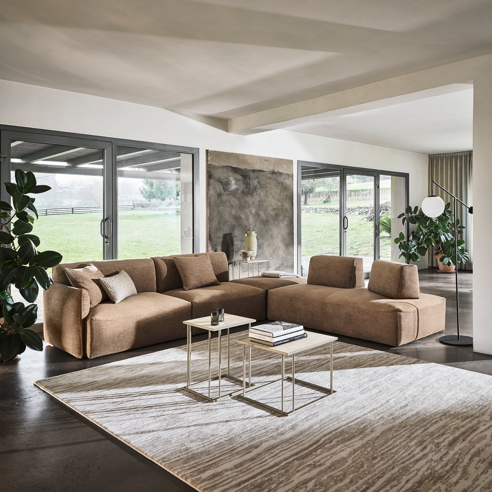 A modern sectional sofa in a warm taupe-brown color arranged on a large textured area rug in a contemporary open-plan living room with large windows and views of a green lawn.