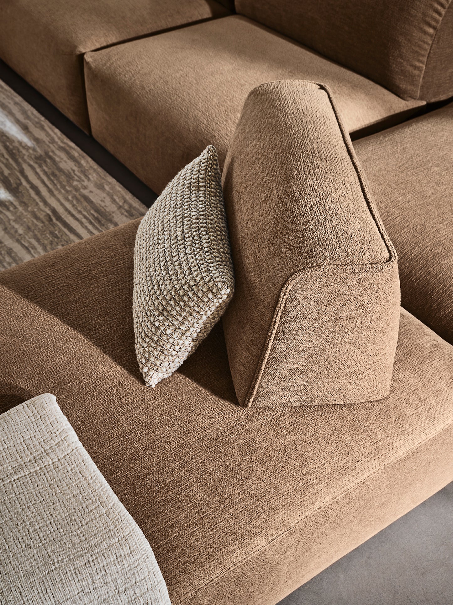 A close-up of a modern sectional sofa in a warm taupe-brown textured fabric, styled with two accent pillows including one chunky knit throw pillow.