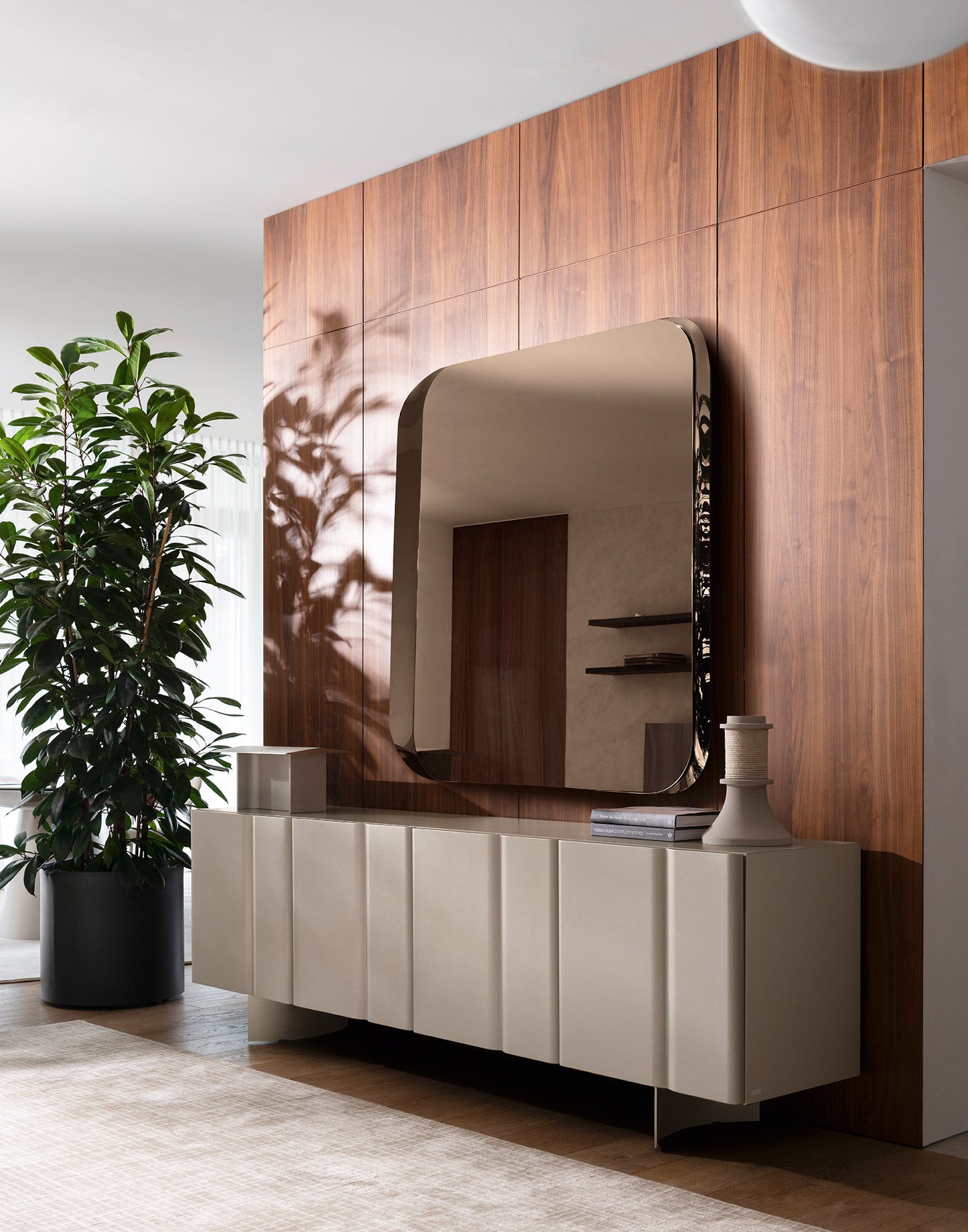 A modern sideboard in a warm beige tone with a large arched mirror mounted above it, set against a wood-paneled wall in a styled interior. A potted plant and decorative objects are visible on and around the cabinet.