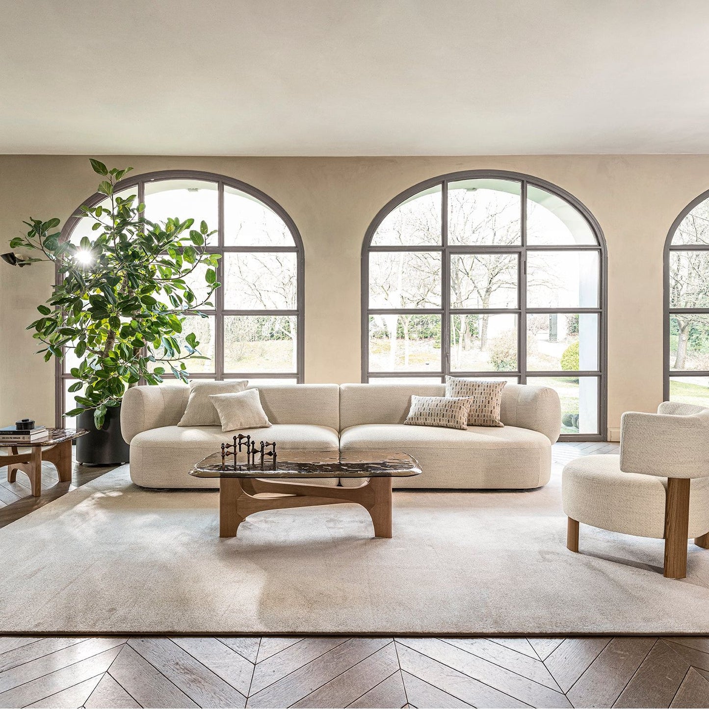 A styled living room interior featuring a cream boucle sofa, a matching armchair, a wooden coffee table, and a large beige area rug, set against arched windows with a potted plant and wood chevron flooring.