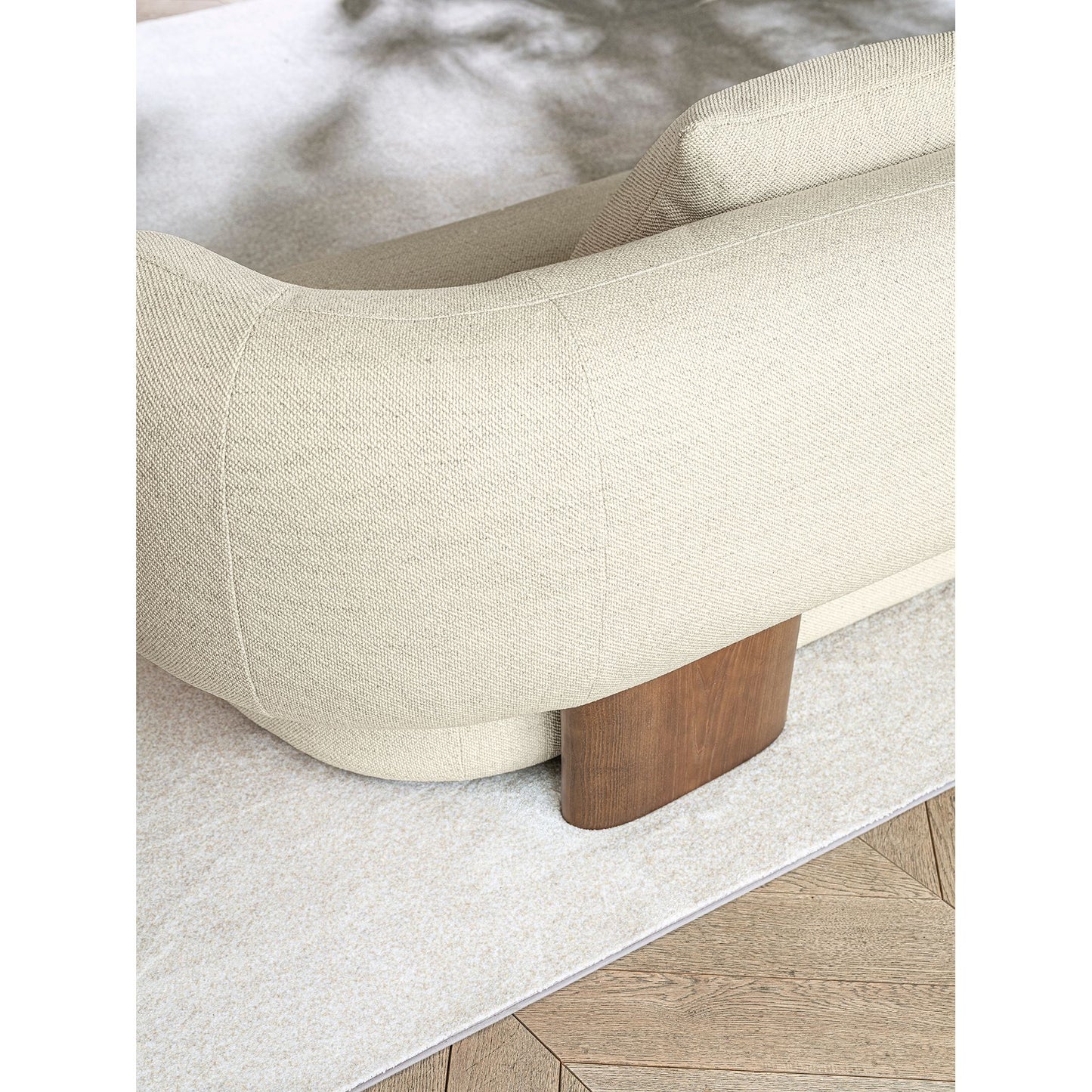 Close-up of a modern upholstered sofa corner in warm off-white textured fabric, showing a curved arm, a wooden leg, and part of a light rug over a wood floor.