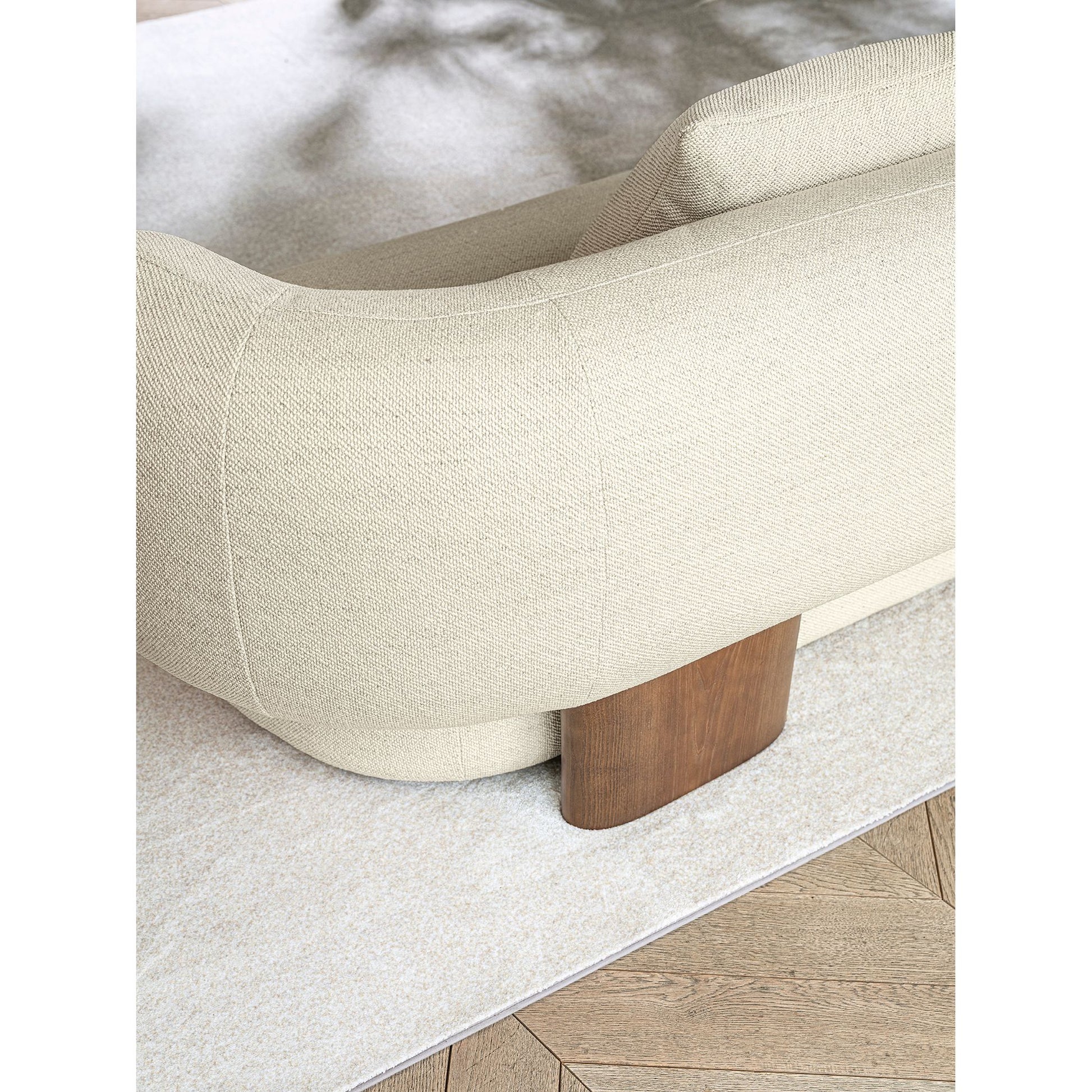 Close-up of a modern upholstered sofa corner in warm off-white textured fabric, showing a curved arm, a wooden leg, and part of a light rug over a wood floor.