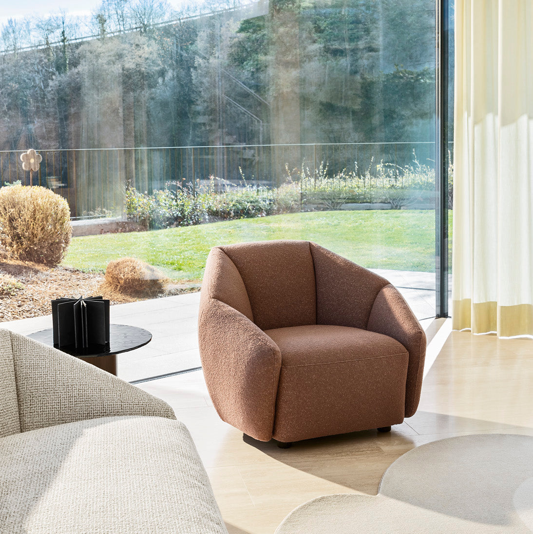 A modern brown upholstered armchair in a bright, contemporary living room with a large floor-to-ceiling window overlooking a landscaped garden. A light beige sofa is partially visible in the foreground, and a round gray rug sits on the floor.