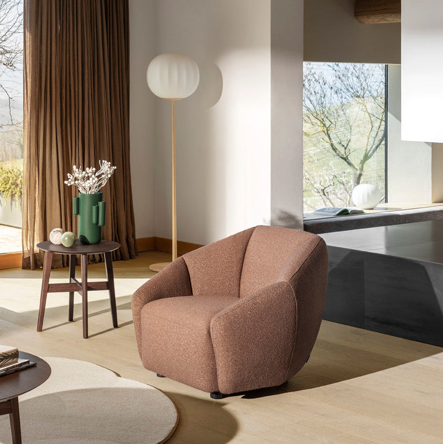 A modern upholstered armchair in a warm taupe-brown fabric, placed in a minimalist, sunlit living room with wooden ceiling beams, a floor lamp, and a side table.
