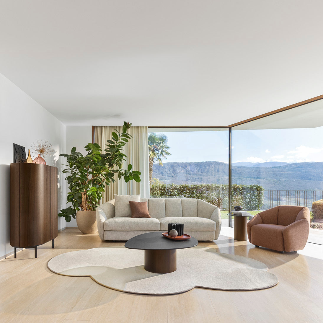 A modern, sunlit living room with light wood floors, white walls, a cream sofa, a brown accent chair, a round black coffee table on a cloud-shaped rug, and large floor-to-ceiling windows overlooking a mountain landscape.