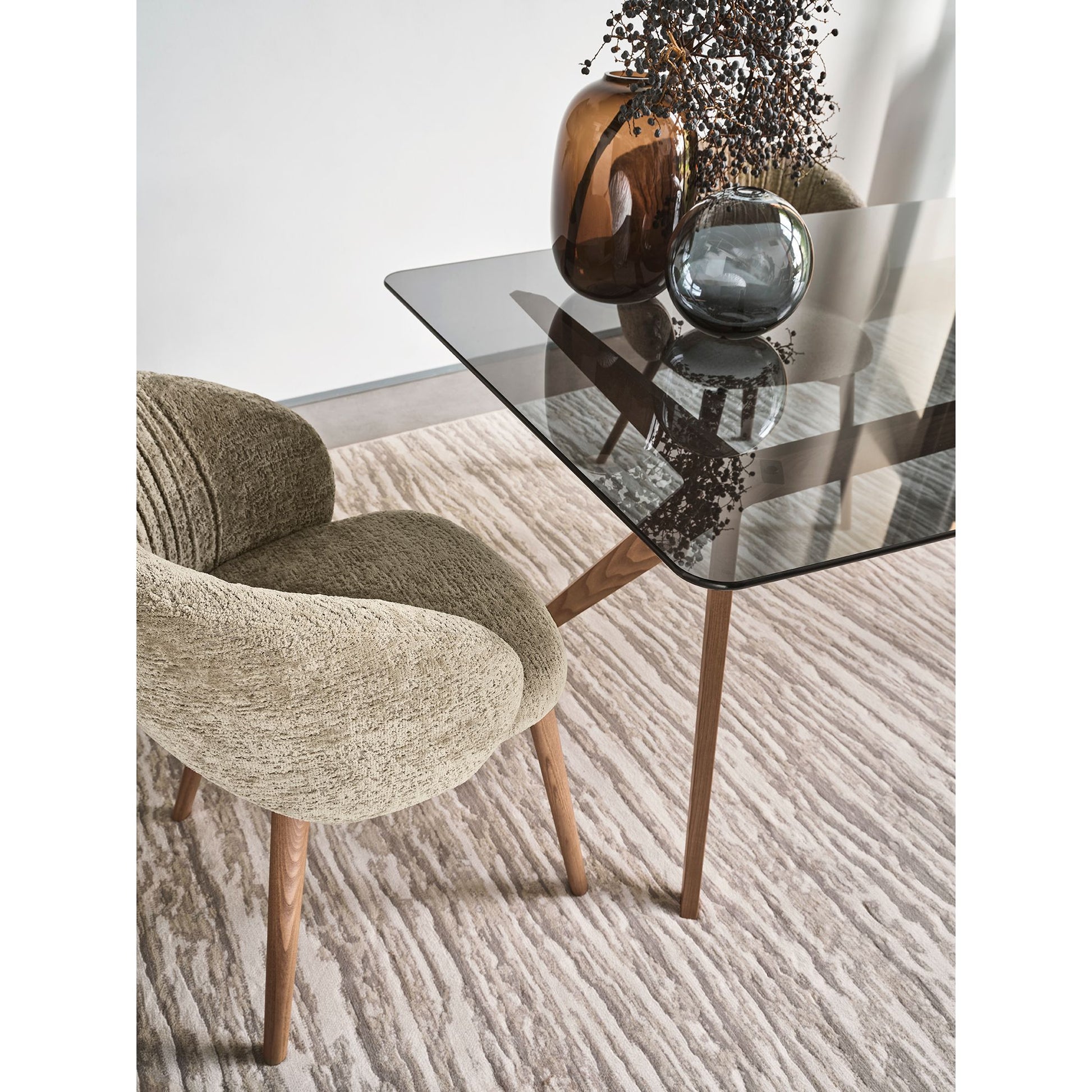 A modern dining setup featuring a textured beige upholstered armchair next to a rectangular glass-top table with wooden legs, set on a patterned beige rug.