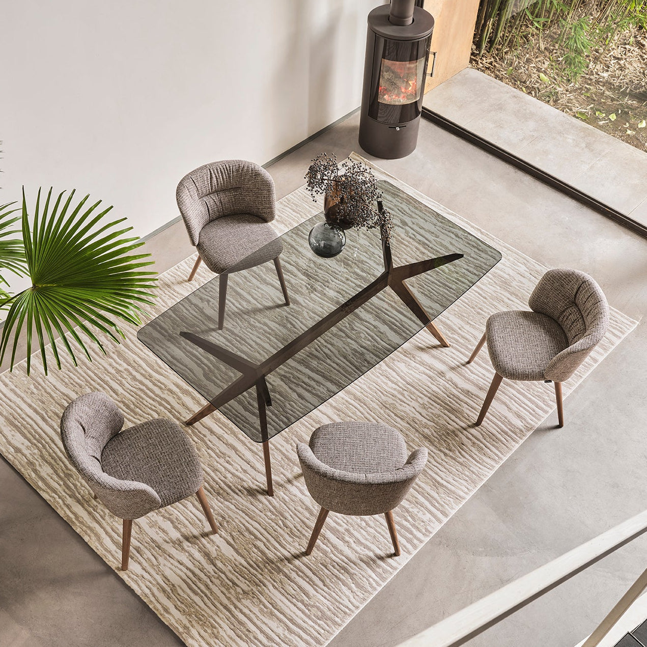 A modern dining room scene viewed from above, featuring four gray upholstered armchairs arranged around a dark wood table on a light rug. Large windows, a glass door, staircase, and indoor plant are visible in the room.