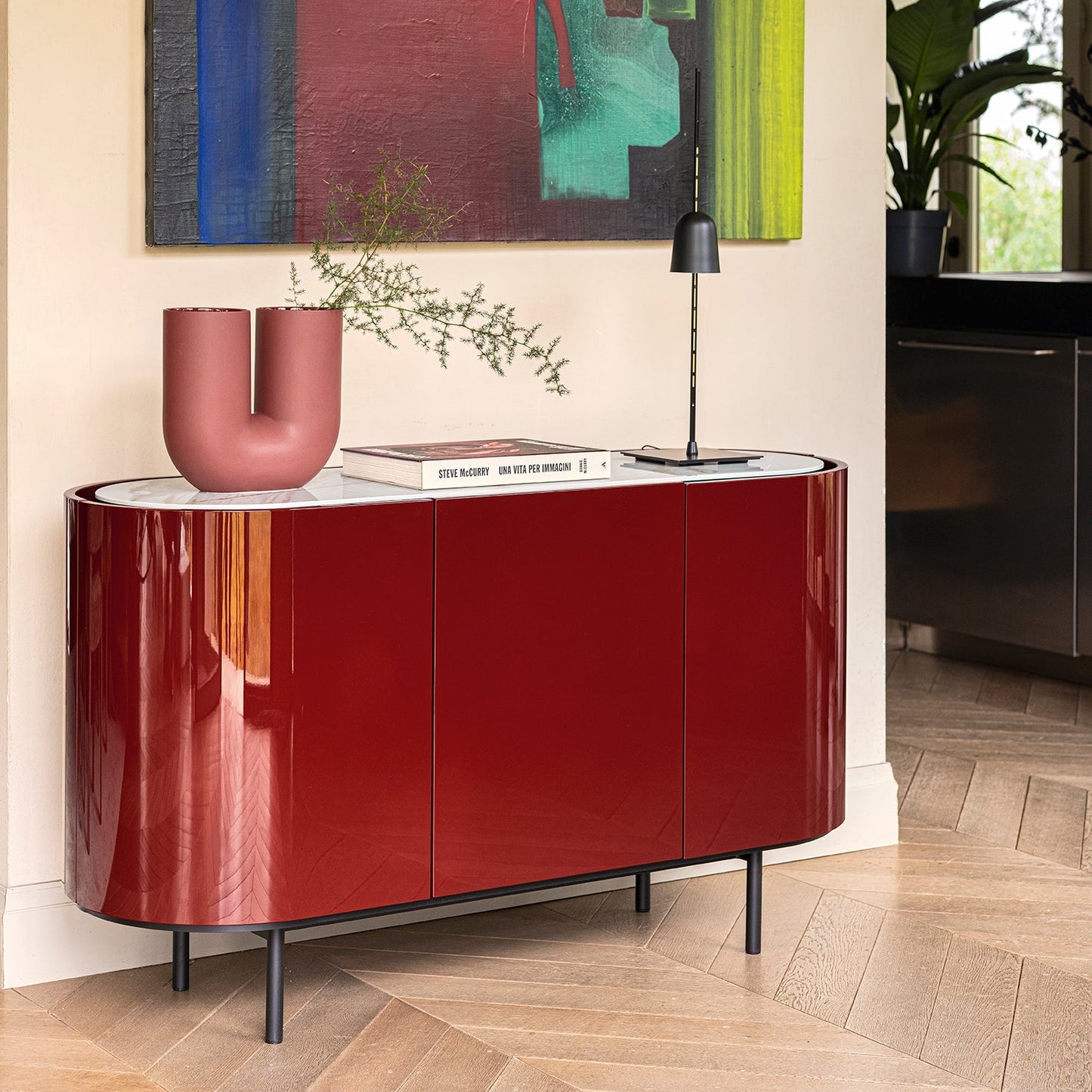 A glossy deep red sideboard cabinet with black legs, styled in a modern entryway with a terracotta vase, books, and a black table lamp. A large abstract painting hangs on the beige wall behind it.
