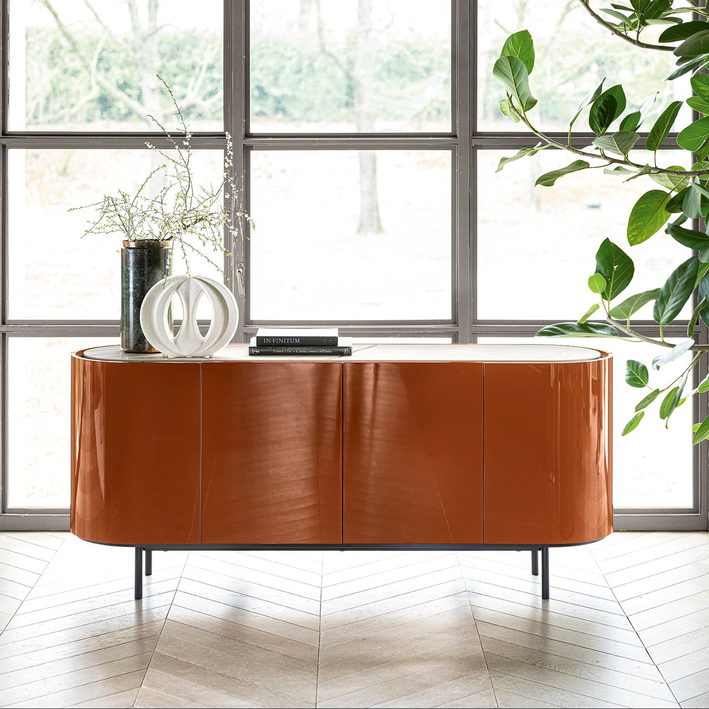 A curved sideboard cabinet in a glossy warm brown finish, styled with decorative objects on top, set in front of a large arched window with a herringbone wood floor and a green plant in the foreground.