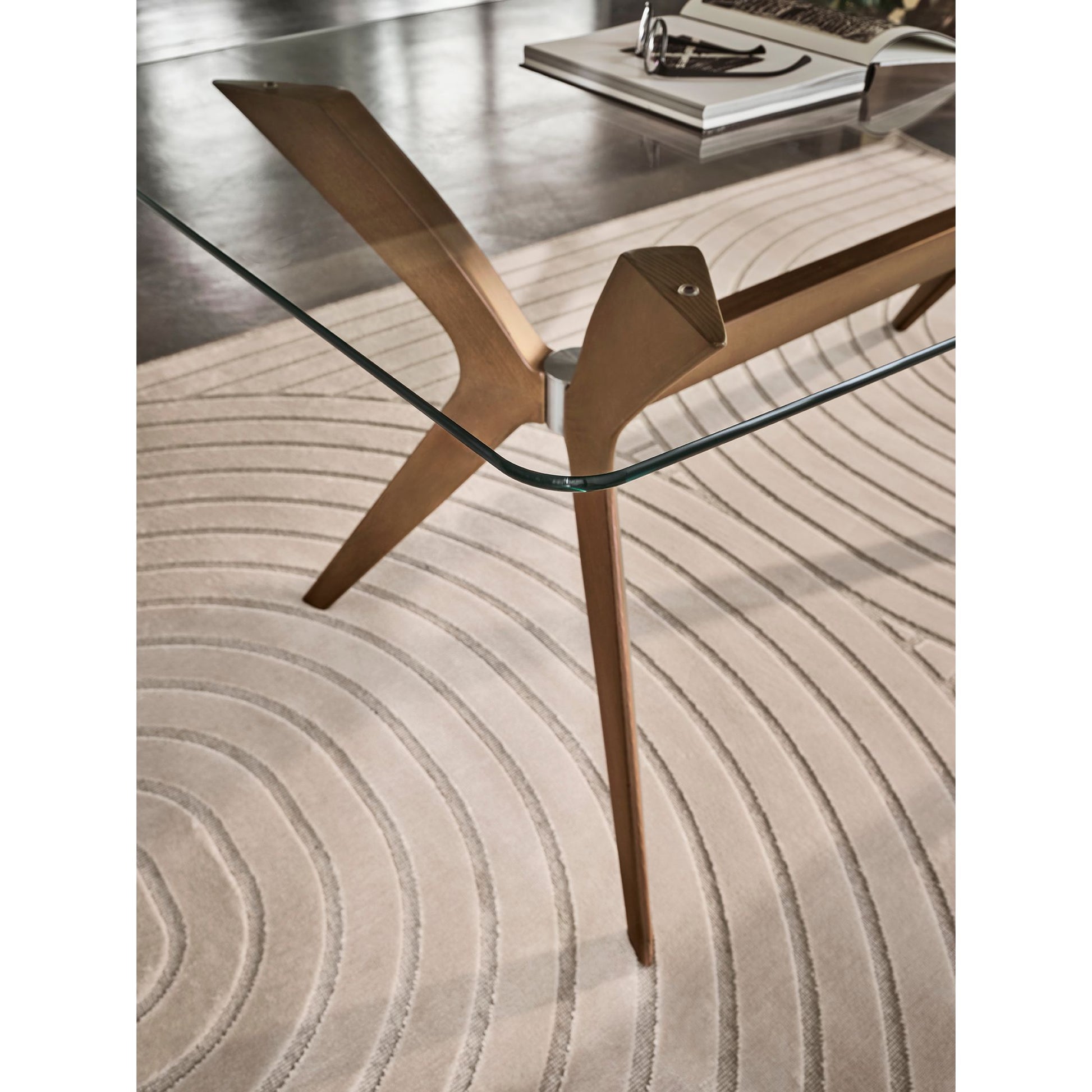 A close-up of a modern glass-top table with a sculptural brass or bronze metal base, resting on a textured beige circular rug.