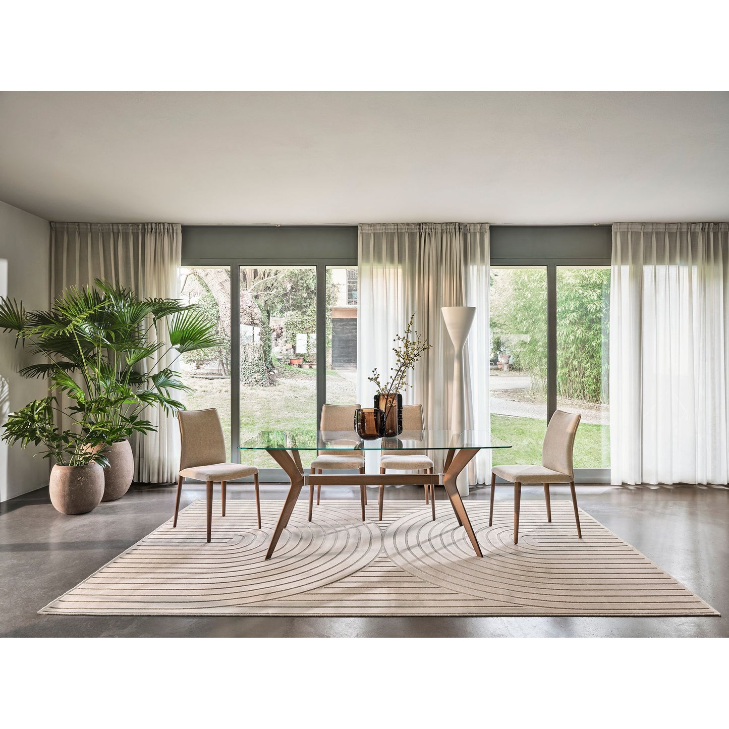 A modern dining room setup featuring a rectangular glass-top dining table with wooden legs, surrounded by four upholstered dining chairs on a large patterned beige rug. The room has large floor-to-ceiling windows with sheer curtains, a potted palm, and a tall floor lamp.