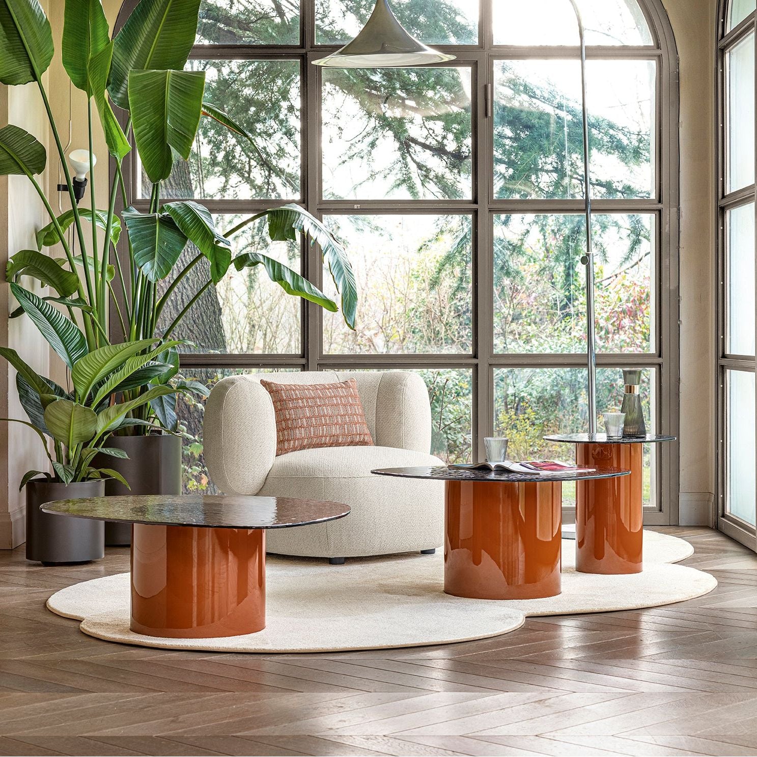 A modern living room scene with a white curved armchair, glossy copper cylindrical coffee tables, a round white rug, large potted plants, and tall multi-pane windows looking out to trees.