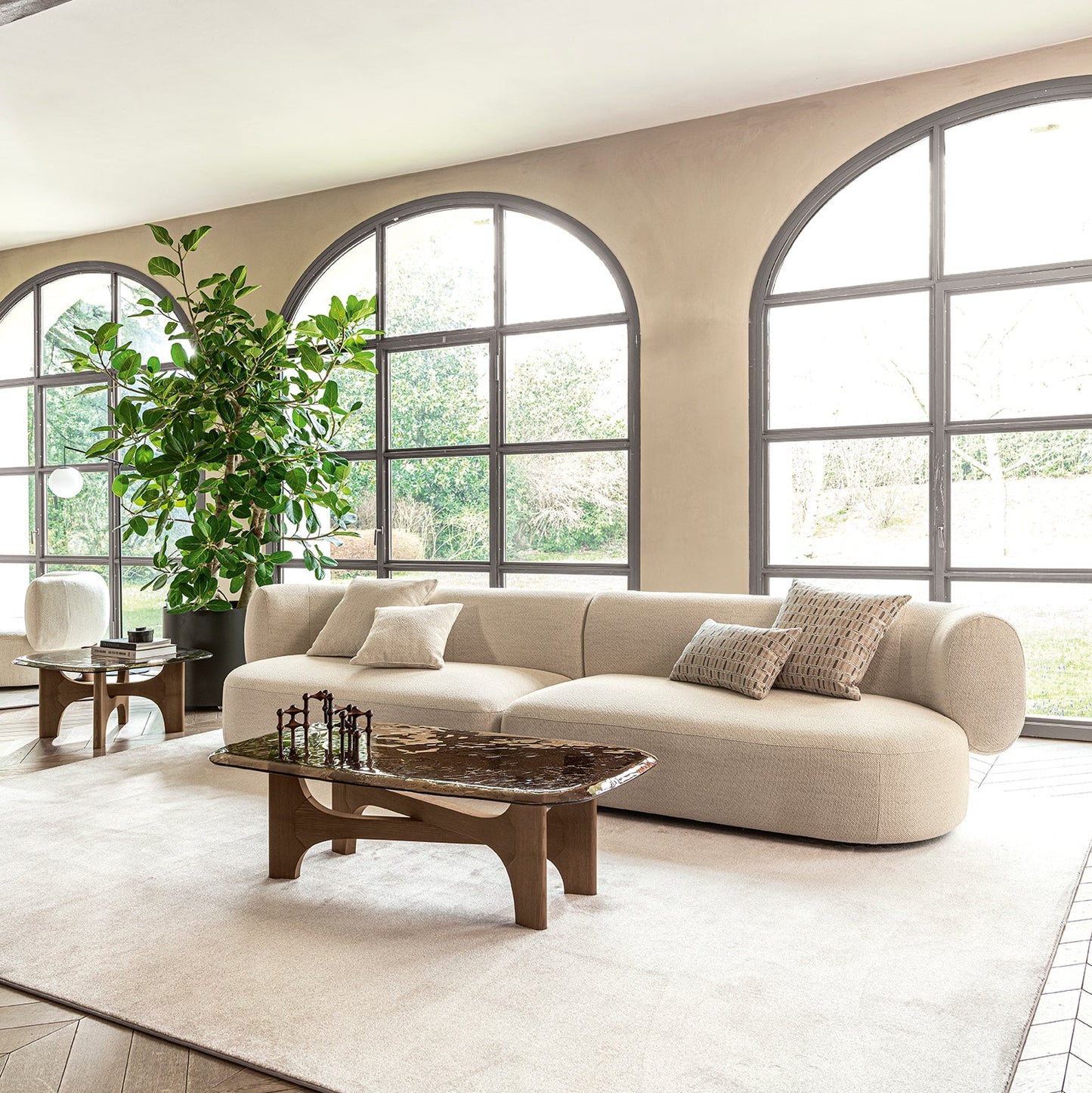 A modern curved beige sofa with neutral throw pillows in a bright, contemporary living room featuring large arched windows, a light rug, and a wooden coffee table.
