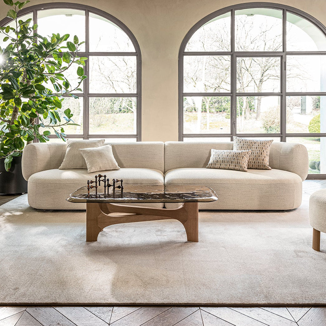 A styled living room interior featuring a cream boucle sofa, a matching armchair, a wooden coffee table, and a large beige area rug, set against arched windows with a potted plant and wood chevron flooring.