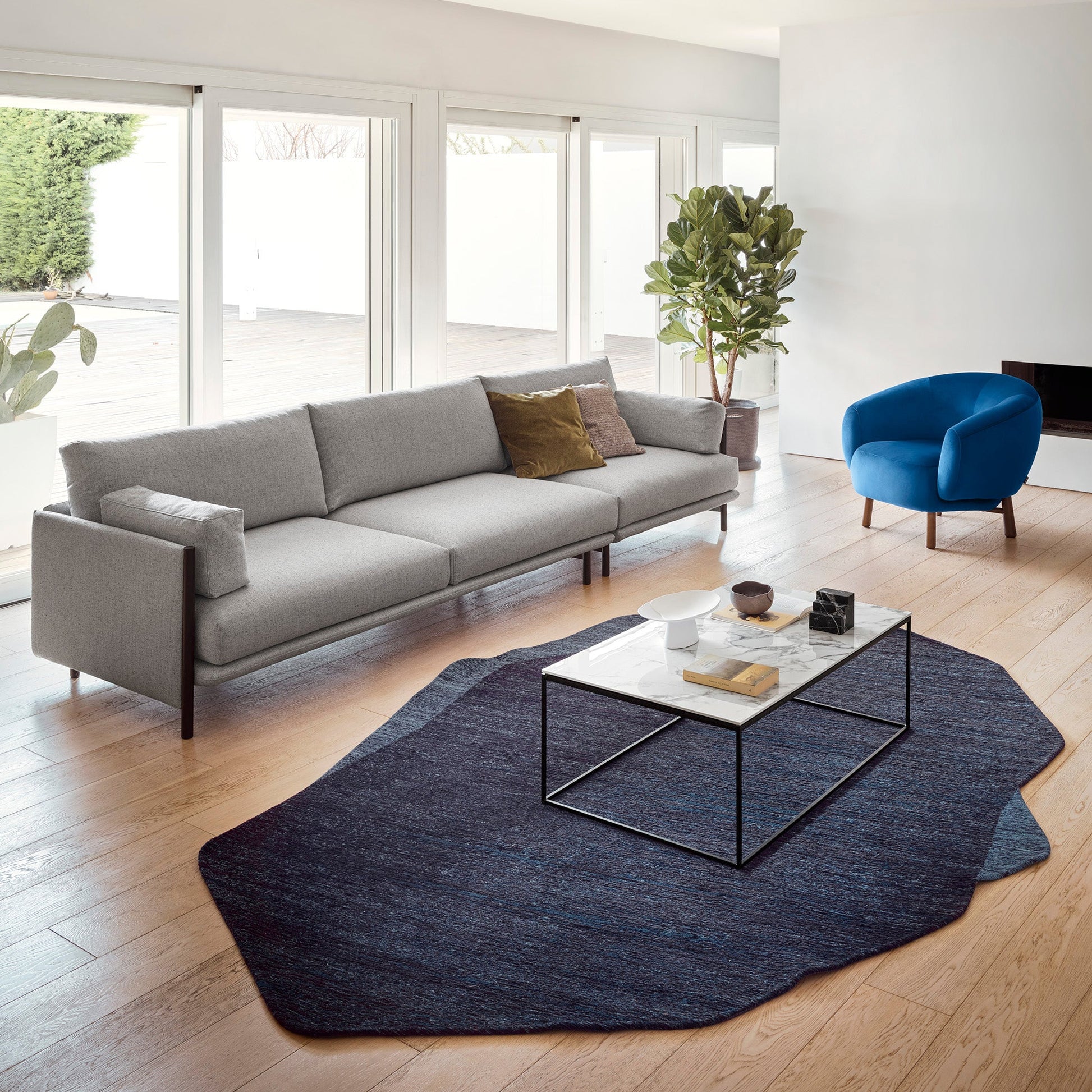 A modern living room featuring a light gray three-seater sofa, a blue accent chair, a dark blue patterned rug, and a glass-top coffee table on a light wood floor.