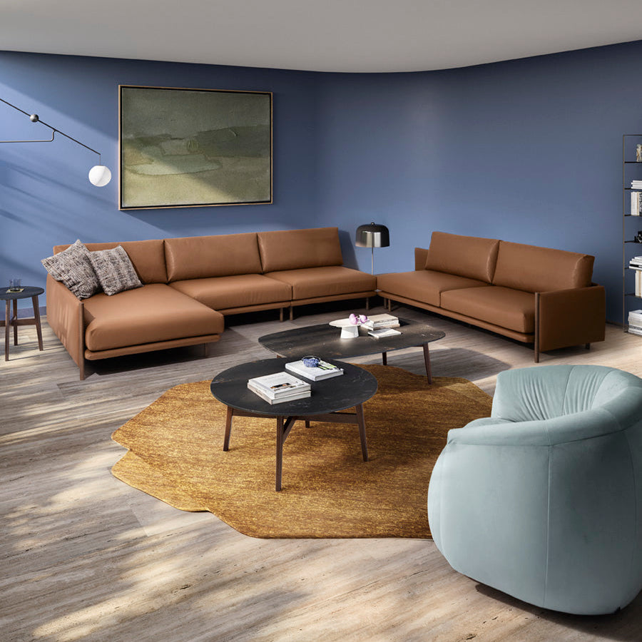 A modern living room setup featuring a brown modular sectional sofa, a light blue armchair, round black coffee tables, and a textured yellow rug on a gray wood floor against a blue wall.
